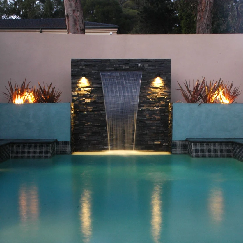 Pool mit Wasserfall umrandet mit schwarzem Stein im dunken beleuchtet..